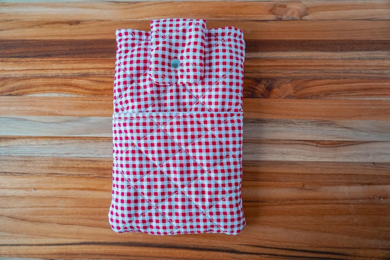 May include: A red and white gingham quilted fabric case with a button closure. The case has a diamond pattern and is designed to hold a tablet or similar device. The case is on a wooden surface.