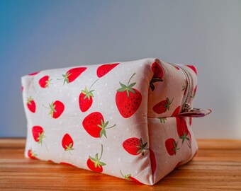 May include: A rectangular, beige fabric cosmetic bag with a repeating pattern of red strawberries and green leaves. The bag has a silver zipper closure and a small pull tab. The background is a gradient of blue and white.