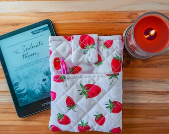 May include: A quilted fabric sleeve featuring a red strawberry pattern on a white background with a snap closure flap and a front pocket containing a pink pen. Next to it is a black e-reader displaying The Soulmate Theory and a lit candle.