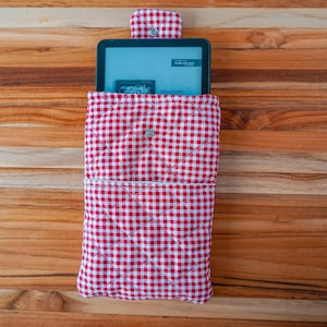 May include: A red and white gingham-patterned tablet sleeve with a button closure. The sleeve is quilted and holds a tablet with a screen displaying text. The background is a wooden surface.