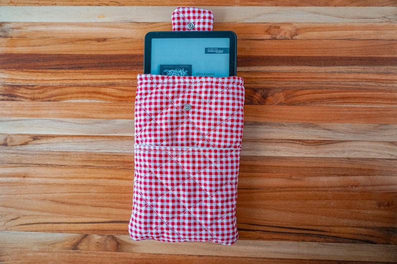 May include: A red and white gingham-patterned tablet sleeve with a button closure. The sleeve is quilted and holds a tablet with a screen displaying text. The background is a wooden surface.