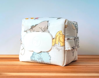 May include: A square, fabric wipe container featuring a Winnie the Pooh design. The container is white with a repeating pattern of the characters. It has a white plastic lid for easy access to wipes. The container is sitting on a wooden surface.