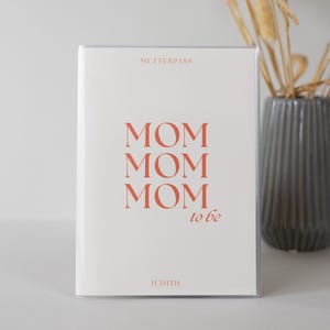 Könnte beinhalten: Ein weißer rechteckiger Dokumentenhalter mit einer transparenten Kunststoffhülle. Der Text MUTTERPASS ist oben in Orange gedruckt. Darunter steht das Wort MOM dreimal in Orange, gefolgt von dem Text to be. Der Name JUDITH steht unten.