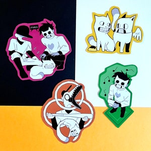 Op de afbeelding: Vijf kleurrijke stickers met cartoonfiguren. De stickers zijn op een witte en zwarte achtergrond. Eén sticker toont een personage met een honkbalknuppel en een kat. Een andere sticker toont een personage met een kat en de tekst "miaou". Een andere sticker toont een personage met een kat. Een andere sticker toont een personage met een kat. Een andere sticker toont een personage met een kat.