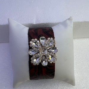 Pu&ograve; includere: Un braccialetto in pelle marrone con un grande fiore di strass trasparente.