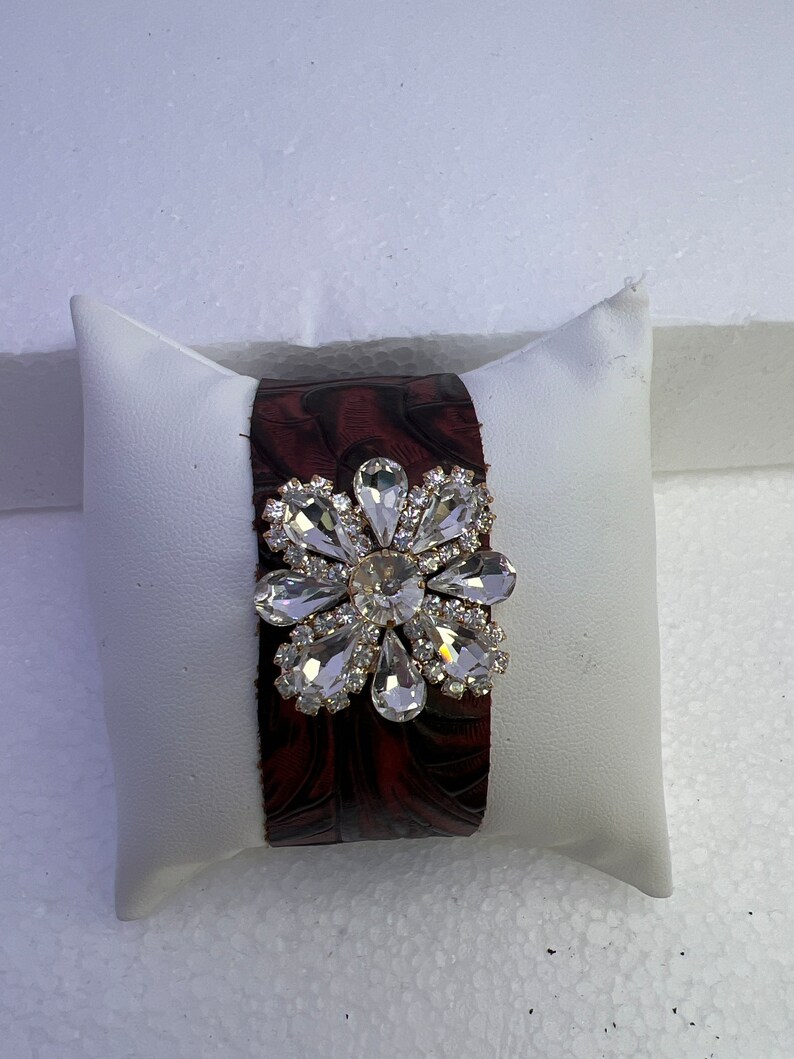 Pu&ograve; includere: Un braccialetto in pelle marrone con un grande fiore di strass trasparente.