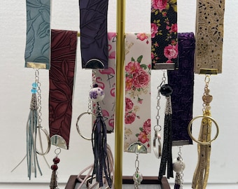 May include: A display of eight keychains with different colored tassels and charms. The keychains are hanging from a gold metal stand. The keychains are made of leather, fabric, and metal. The tassels are made of leather, fabric, and beads. The charms are made of metal, ceramic, and glass.
