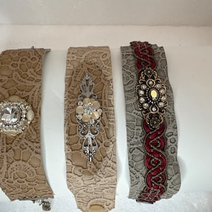 May include: Three leather bracelets with different embellishments. The first bracelet has a clear rhinestone and pearl accents. The second bracelet has a silver floral design with a pearl centre. The third bracelet has a red braided cord with a gold and rhinestone embellishment.