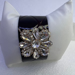 Pu&ograve; includere: Un braccialetto rigido in pelle trapuntata nera con un grande fiore di strass trasparente.