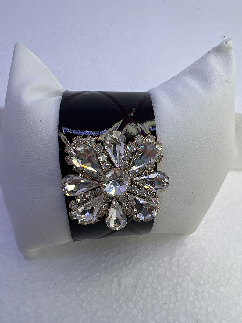 Pu&ograve; includere: Un braccialetto rigido in pelle trapuntata nera con un grande fiore di strass trasparente.