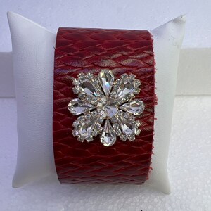 Pu&ograve; includere: Un braccialetto in pelle rossa con un grande decoro floreale in strass trasparente.
