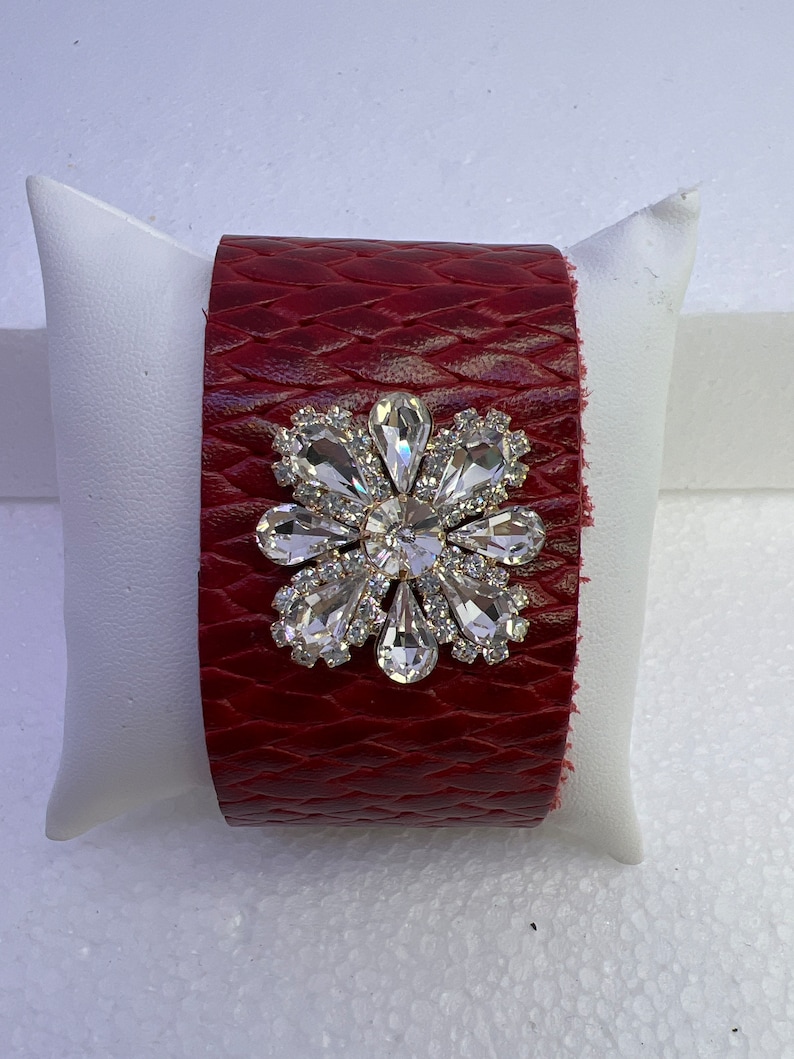 Pu&ograve; includere: Un braccialetto in pelle rossa con un grande decoro floreale in strass trasparente.