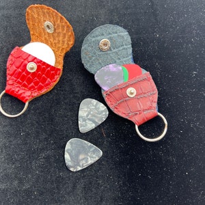 May include: Two red leather guitar pick holders with keychains. One holder is open and shows a white pick inside. The other holder is closed and has three picks inside. Two additional picks are on the black surface.