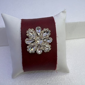 Pu&ograve; includere: Un braccialetto in pelle bordeaux con un grande fiore di strass trasparente.