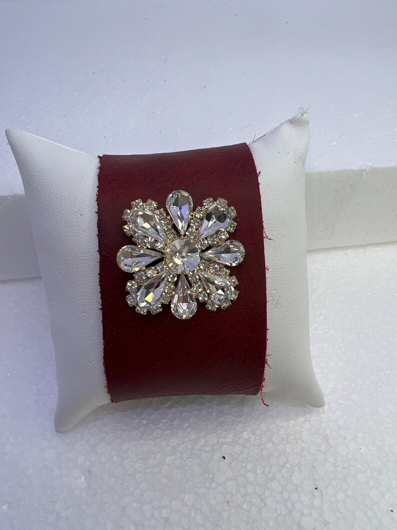 Pu&ograve; includere: Un braccialetto in pelle bordeaux con un grande fiore di strass trasparente.