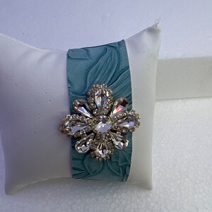 Pu&ograve; includere: Un braccialetto in pelle blu-verde con un grande spilla a forma di fiore di cristallo trasparente.
