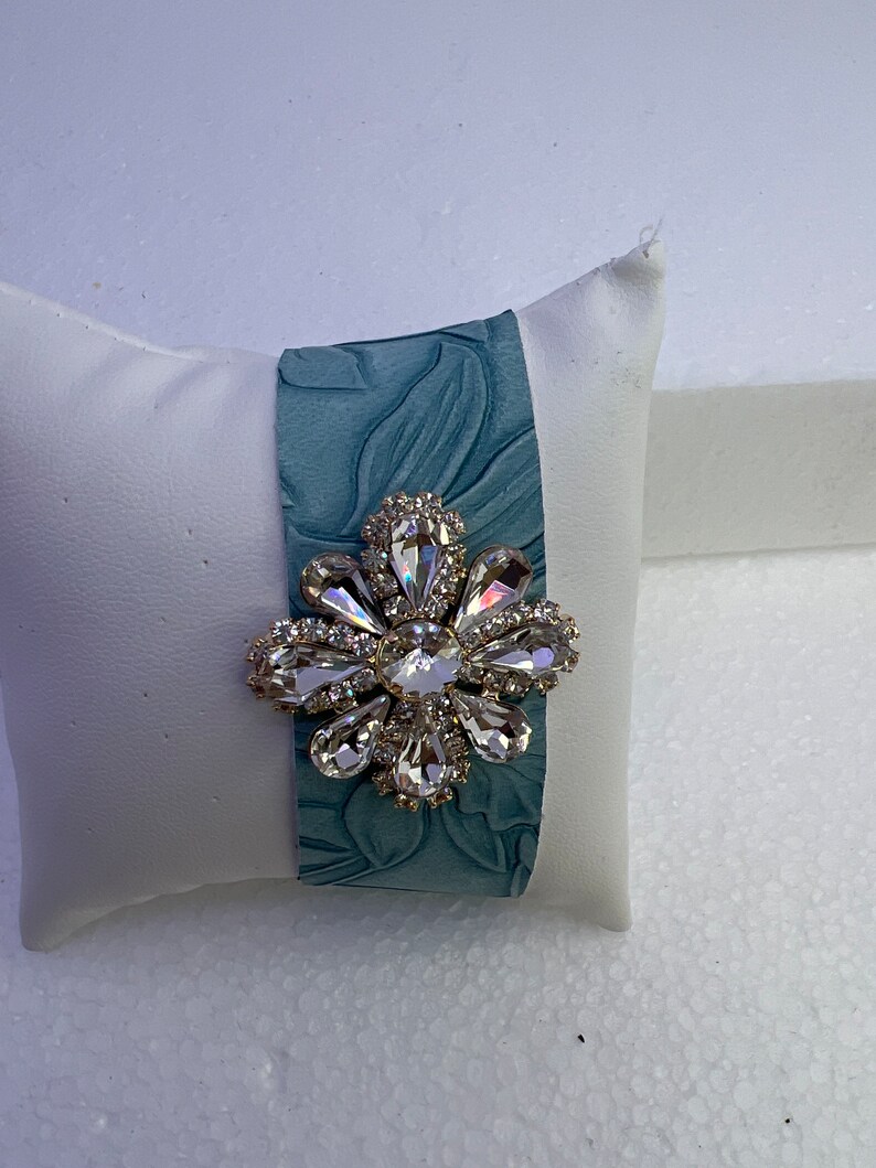 Pu&ograve; includere: Un braccialetto in pelle blu-verde con un grande spilla a forma di fiore di cristallo trasparente.