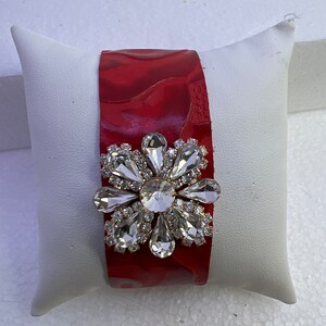 Pu&ograve; includere: Un braccialetto in pelle rossa con un grande fiore di strass trasparente.