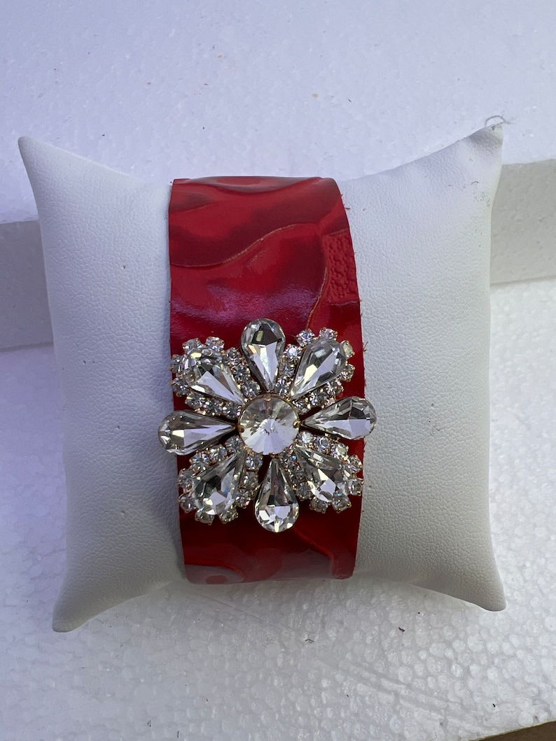 Pu&ograve; includere: Un braccialetto in pelle rossa con un grande fiore di strass trasparente.