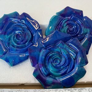 May include: Three blue and purple ceramic rose wall decor pieces. The roses are stylised and have a swirl pattern in the centre.