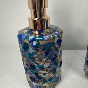 May include: A blue and gold soap dispenser with a geometric design. The dispenser is made of clear resin with embedded pieces of seashells and glass.