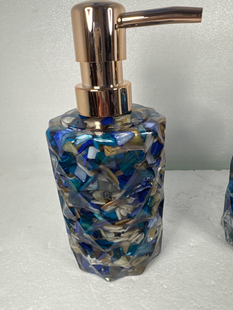 May include: A blue and gold soap dispenser with a geometric design. The dispenser is made of clear resin with embedded pieces of seashells and glass.