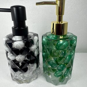 May include: Two decorative soap dispensers with clear glass bottles filled with colorful stones. The dispenser on the left has black and white stones, while the dispenser on the right has green stones.