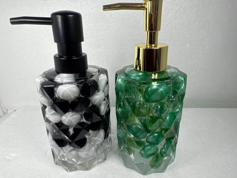 May include: Two decorative soap dispensers with clear glass bottles filled with colorful stones. The dispenser on the left has black and white stones, while the dispenser on the right has green stones.