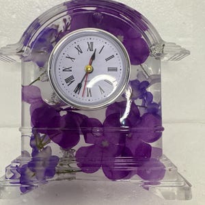 May include: A clear resin clock with a white clock face and black Roman numerals. The clock is adorned with purple flowers, creating a unique decorative piece. The clock measures approximately 5 inches tall.