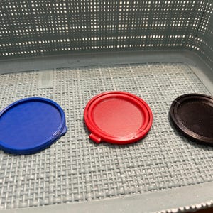 May include: Three circular, 3D-printed lids in blue, red, and black. The lids are arranged on a grey, textured surface inside a light blue basket. The lids have a raised edge and a flat top.