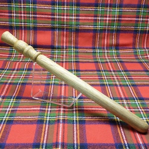 Peut inclure: Une baguette en bois avec une extrémité arrondie et un sommet légèrement évasé. La baguette est de couleur brun clair et repose sur un tissu à carreaux rouge et vert.