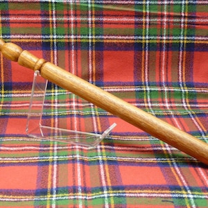 May include: A wooden rolling pin with a brown finish and a slightly rounded handle. The rolling pin is resting on a red and green tartan fabric.