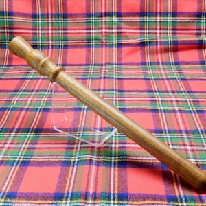 Può includere: Una bacchetta di legno con finitura marrone chiaro, con un'impugnatura dettagliata e un corpo liscio e affusolato. La bacchetta è esposta su un supporto trasparente su uno sfondo di tessuto a quadri rossi, verdi e blu.