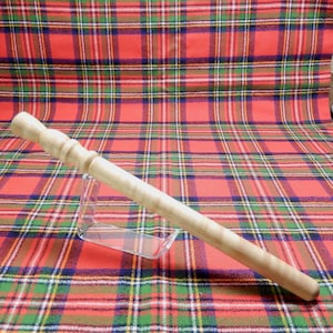 May include: A light-coloured wooden wand with a carved handle and a smooth, cylindrical body. The wand is displayed on a clear acrylic stand against a backdrop of a red, green, and blue plaid pattern.