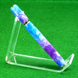 Può includere: Una penna colorata con un design marmorizzato blu, viola e bianco, esposta su un supporto in acrilico trasparente. La penna ha dettagli color oro e un design elegante. Lo sfondo è verde.