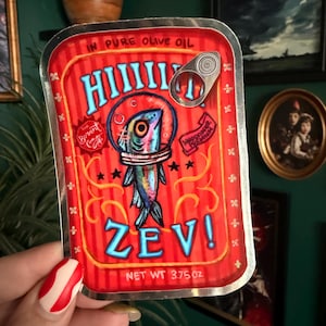 Könnte beinhalten: Ein roter und silberner Aufkleber mit einer Sardine in einem Raumanzug. Der Aufkleber trägt die Aufschrift "HIIIII ZEV!" in blauer und weißer Schrift, mit den Worten "IN PURE OLIVE OIL" oben und "NET WT 106g" unten. Der Aufkleber hat eine Lasche.