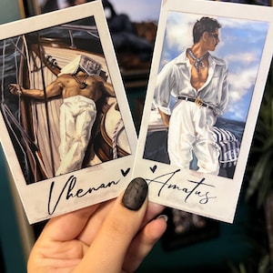 May include: Two polaroid-style prints featuring male figures. One print shows a man on a boat, the other a man in a white shirt. The prints have the names "Vhenan" and "Amatus" written on them in cursive.