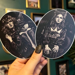 May include: Two oval stickers featuring gothic illustrations in black and white. One depicts a couple embracing, the other a woman holding flowers. The stickers are held in a hand against a background of framed art.