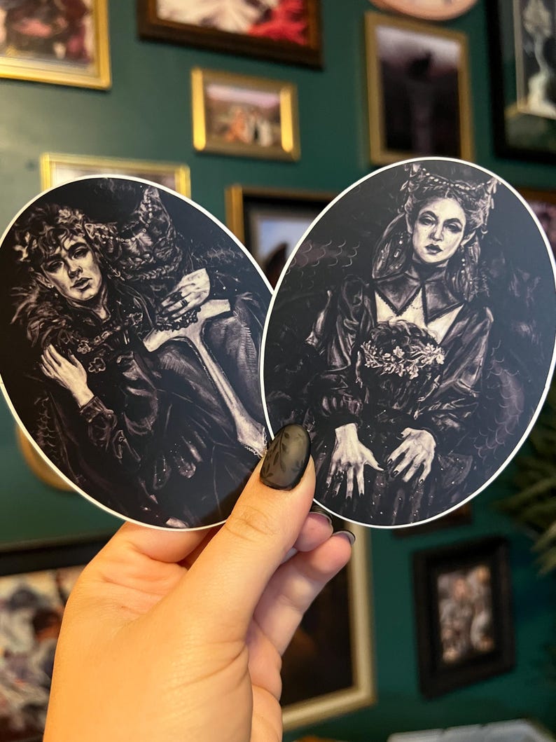 May include: Two oval stickers featuring gothic illustrations in black and white. One depicts a couple embracing, the other a woman holding flowers. The stickers are held in a hand against a background of framed art.
