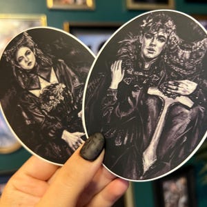 May include: Two oval stickers featuring black and white illustrations of figures in ornate clothing. One sticker depicts a woman with a crown and the other a man with a crown and a scepter. The stickers have a gothic or fantasy theme.