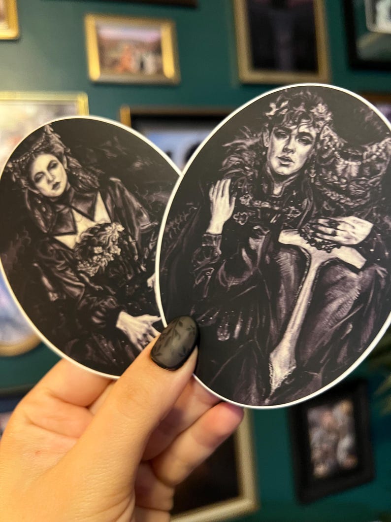 May include: Two oval stickers featuring black and white illustrations of figures in ornate clothing. One sticker depicts a woman with a crown and the other a man with a crown and a scepter. The stickers have a gothic or fantasy theme.