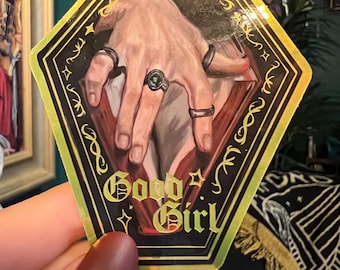 Könnte beinhalten: Ein goldumrandeter Aufkleber mit einer Handillustration, die Ringe trägt, vor dunklem Hintergrund. Der Text "Good Girl" ist in einer dekorativen Schriftart gehalten. Der Aufkleber hat eine sechseckige Form.