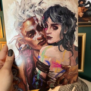 May include: An art print featuring a close embrace between two figures. One figure has white hair and the other has dark hair. The artwork has a painterly style with a palette of browns, pinks, and blues. The print is held by a hand with a black nail.