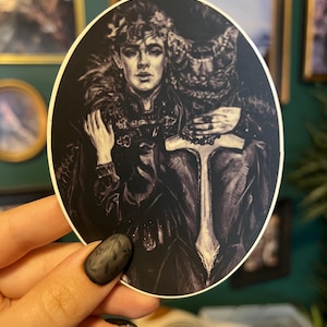 May include: A black and white sticker featuring a detailed illustration of two figures. One figure is a young person with curly hair and a feathered collar. The other figure is partially obscured, with a textured design. The sticker is held by a hand with a black floral nail design.