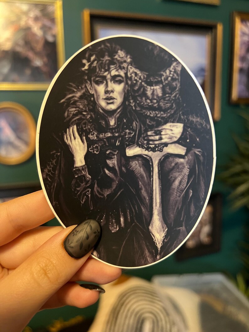 May include: A black and white sticker featuring a detailed illustration of two figures. One figure is a young person with curly hair and a feathered collar. The other figure is partially obscured, with a textured design. The sticker is held by a hand with a black floral nail design.