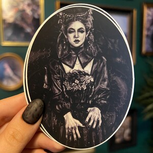 May include: Oval sticker featuring a black and white illustration of a woman in a dark, gothic style dress and crown. The woman has long hair and is holding a bouquet of flowers. The sticker has a white border.
