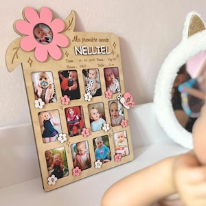 Peut inclure: Cadre photo en bois avec un design de fleur et de papillon. Le cadre contient douze petites photos, chacune entourée d'une fleur rose. Le texte "Ma première année NELLIE" est visible en haut, avec la date et le poids du bébé.