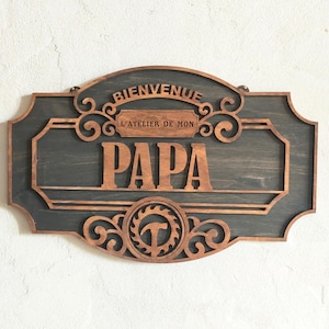 Peut inclure: Une plaque murale rectangulaire en bois aux coins arrondis, avec un fond gris foncé et des découpes en bois clair. Le texte indique BIENVENUE L'ATELIER DE MON PAPA, avec un emblème circulaire en bas représentant une lame de scie et un marteau.