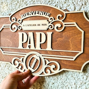Op de afbeelding: Houten bord met de woorden "BIENVENUE A L'ATELIER DE MON PAPI" in een decoratief lettertype. Het bord heeft een voetbalafbeelding en sierlijke krullen. Het bord is gemaakt van gelaagd hout met een donkerbruine vlek en licht houten accenten.
