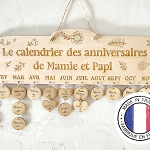 Peut inclure: Un calendrier en bois avec un fond blanc et une bordure brune. Le calendrier est décoré de motifs floraux et du texte "Le calendrier des anniversaires de Mamie et Papi". Le calendrier comporte 12 mois, chacun avec un petit cercle pour un nom et un numéro. Les cercles sont attachés au calendrier avec de petits anneaux métalliques. Le calendrier comporte également un petit cercle avec le texte "MADE IN FRANCE" et un drapeau français.